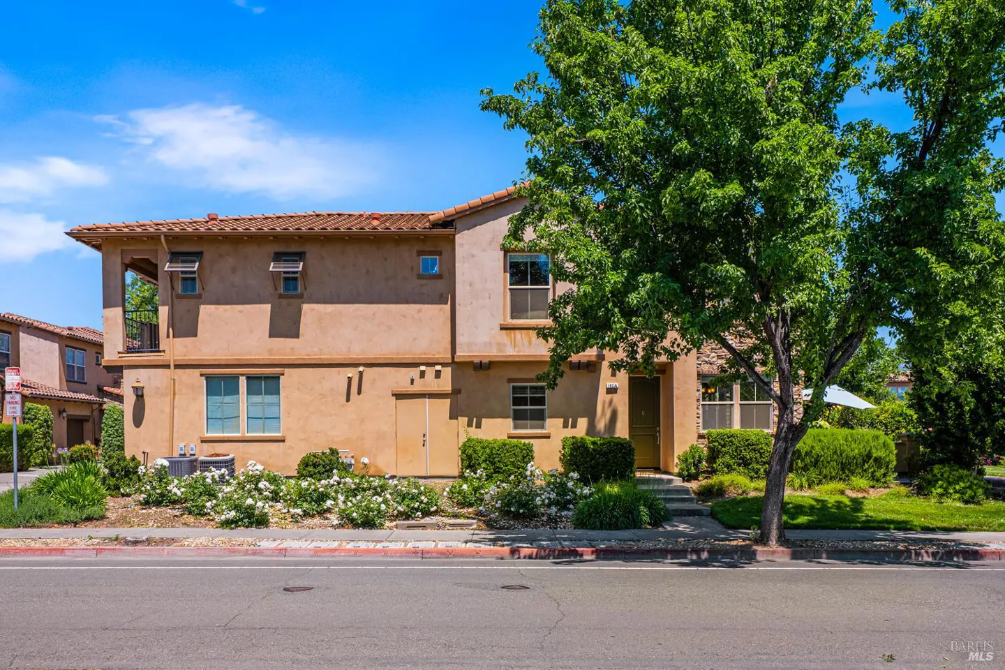 Two-story tan stucco townhouse with a tile roof, surrounded by green trees and white flowering bushes under a blue sky.