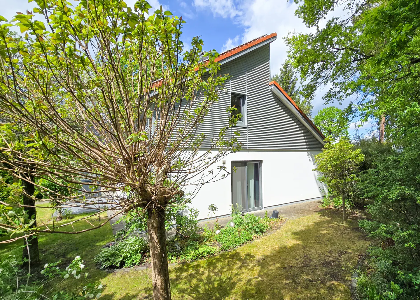 Modern two-story house with grey corrugated siding and white walls, surrounded by a lush green garden and trees.