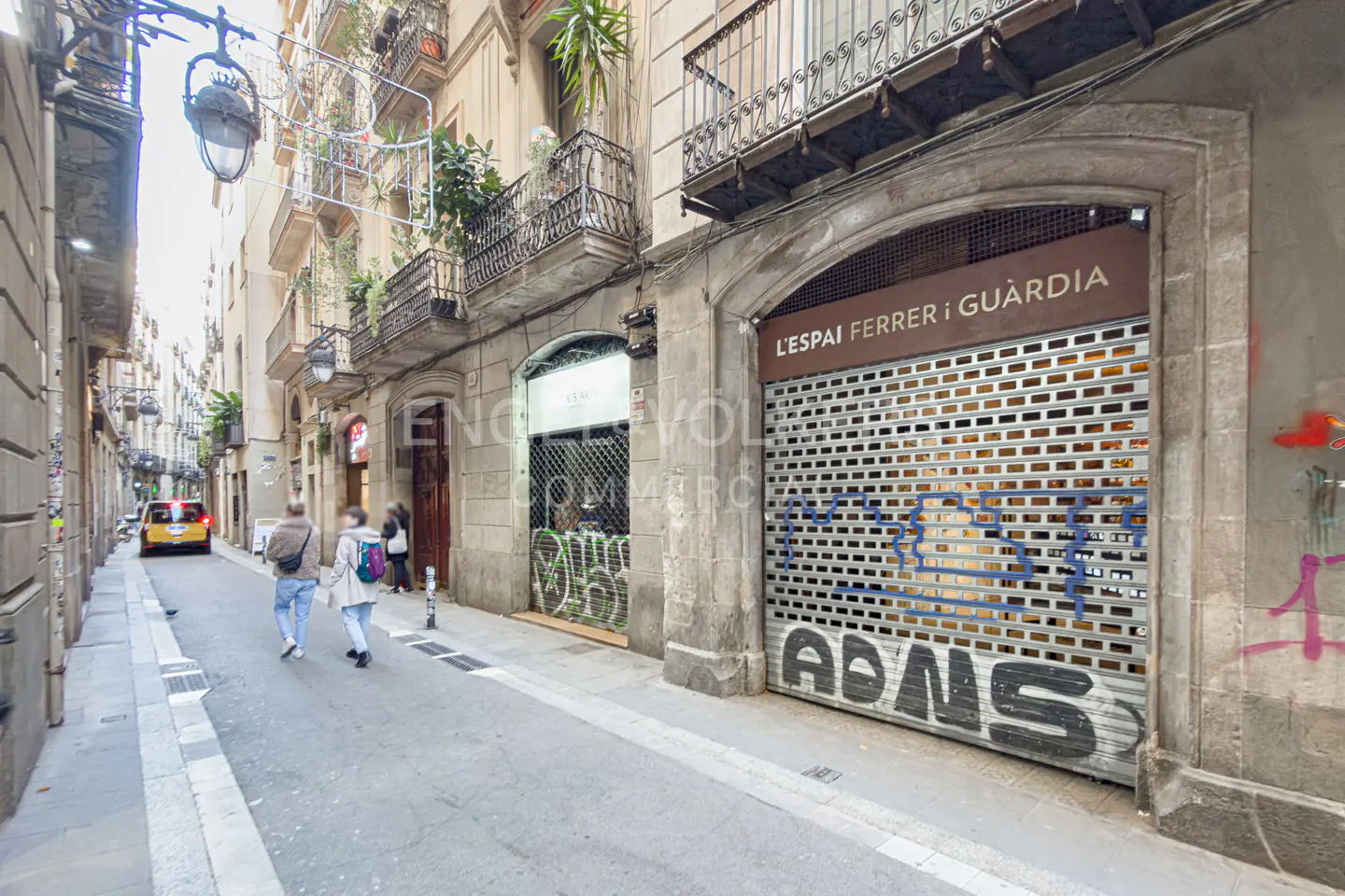 Street view of a narrow road with people walking. Buildings have balconies and shops with metal shutters, some with graffiti.