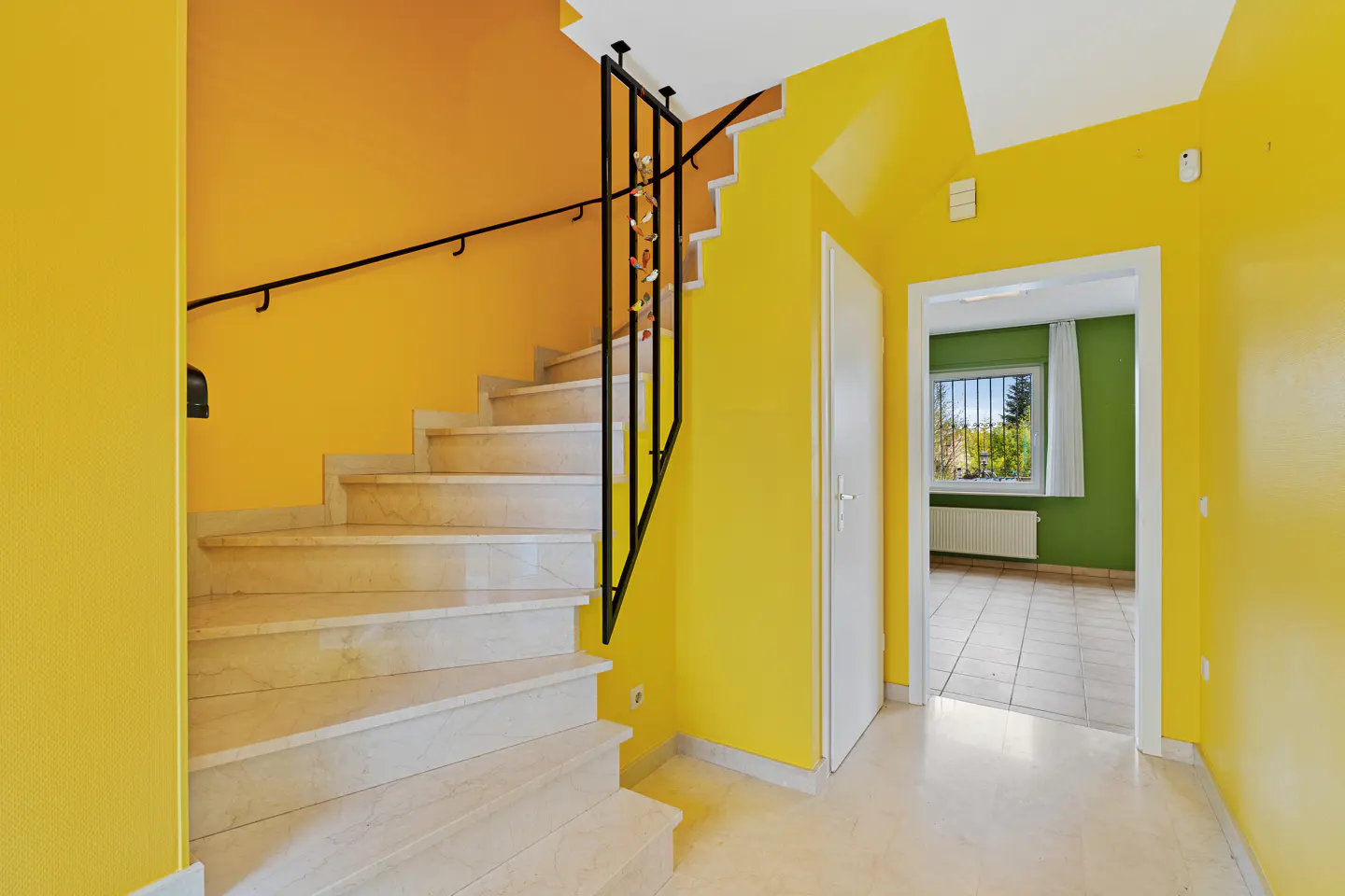 Interior view of a yellow hallway with marble stairs, a black railing, and a doorway to a green room with a window.