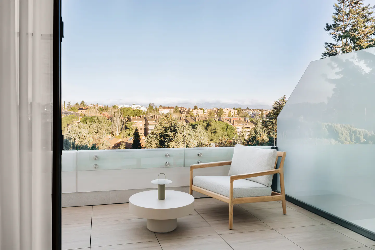 Balcony view with a white cushioned chair, round table with a green lamp, glass railing, and a city view in the background.