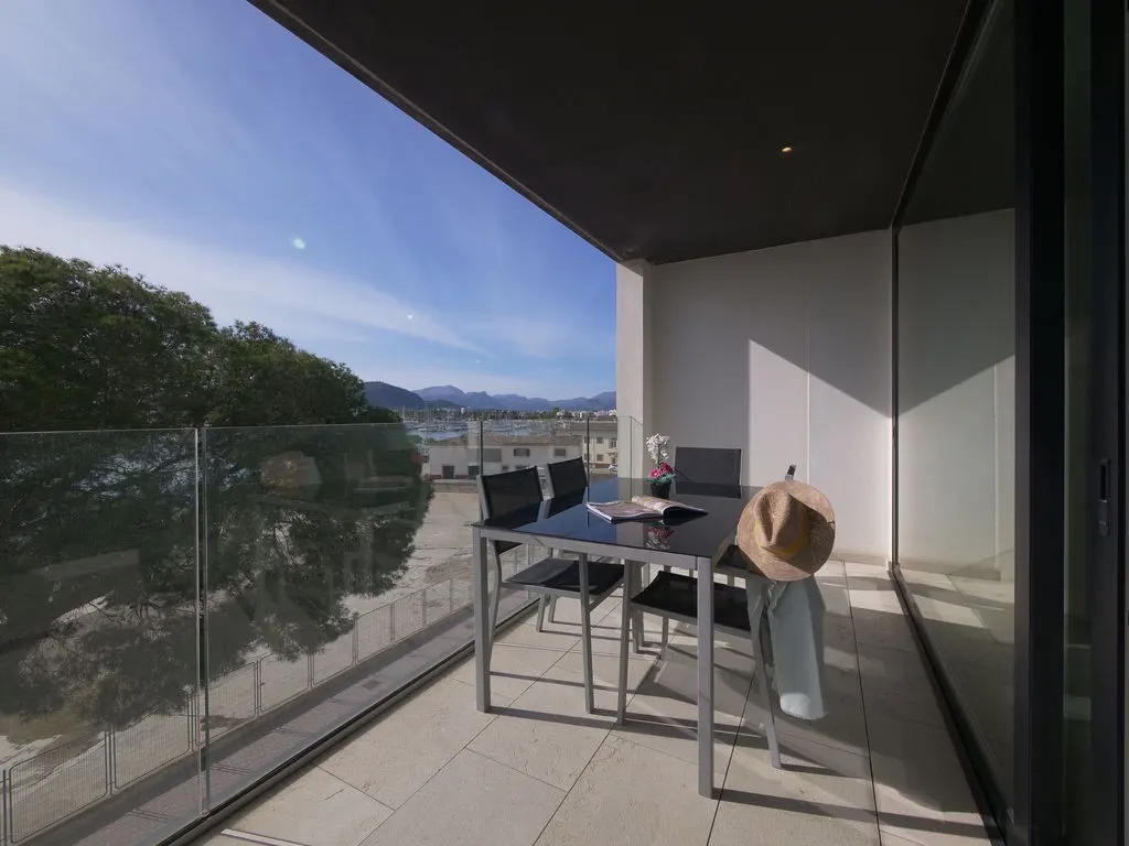Balcony with a glass table, four chairs, a hat, and a view of trees, water, and mountains under a blue sky.