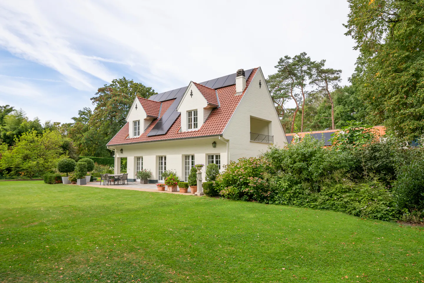 A white house with a red tile roof and solar panels sits on a green lawn surrounded by trees.
