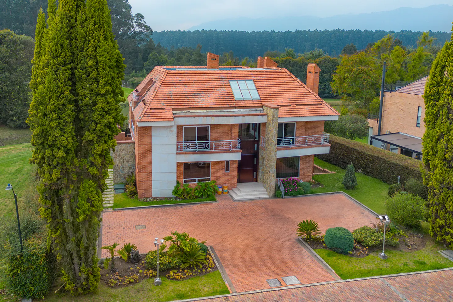 Two-story brick house with a red tile roof, surrounded by green trees and a brick driveway.