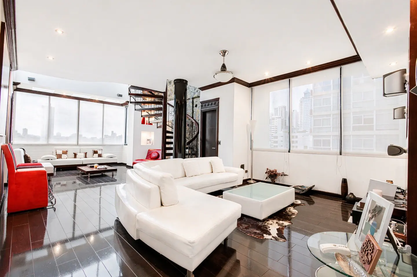 Bright living room with white sofas, dark wood floors, and a spiral staircase. Large windows offer a city view.