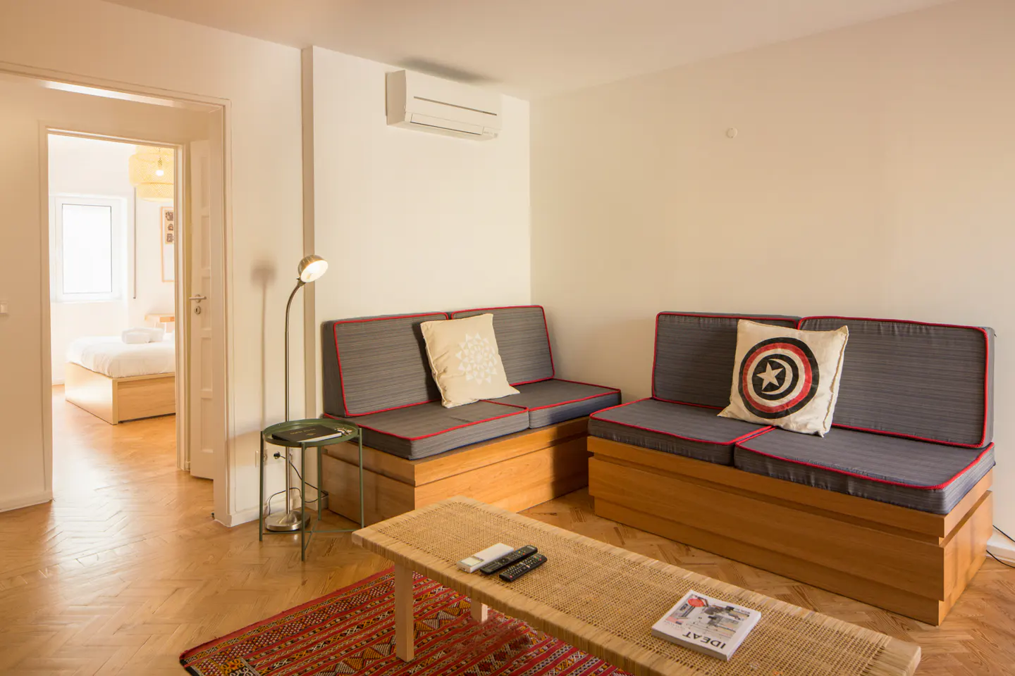 Living room with gray sofas, red trim, wooden base, and Captain America pillow. Woven coffee table and red rug. Bedroom visible through doorway.