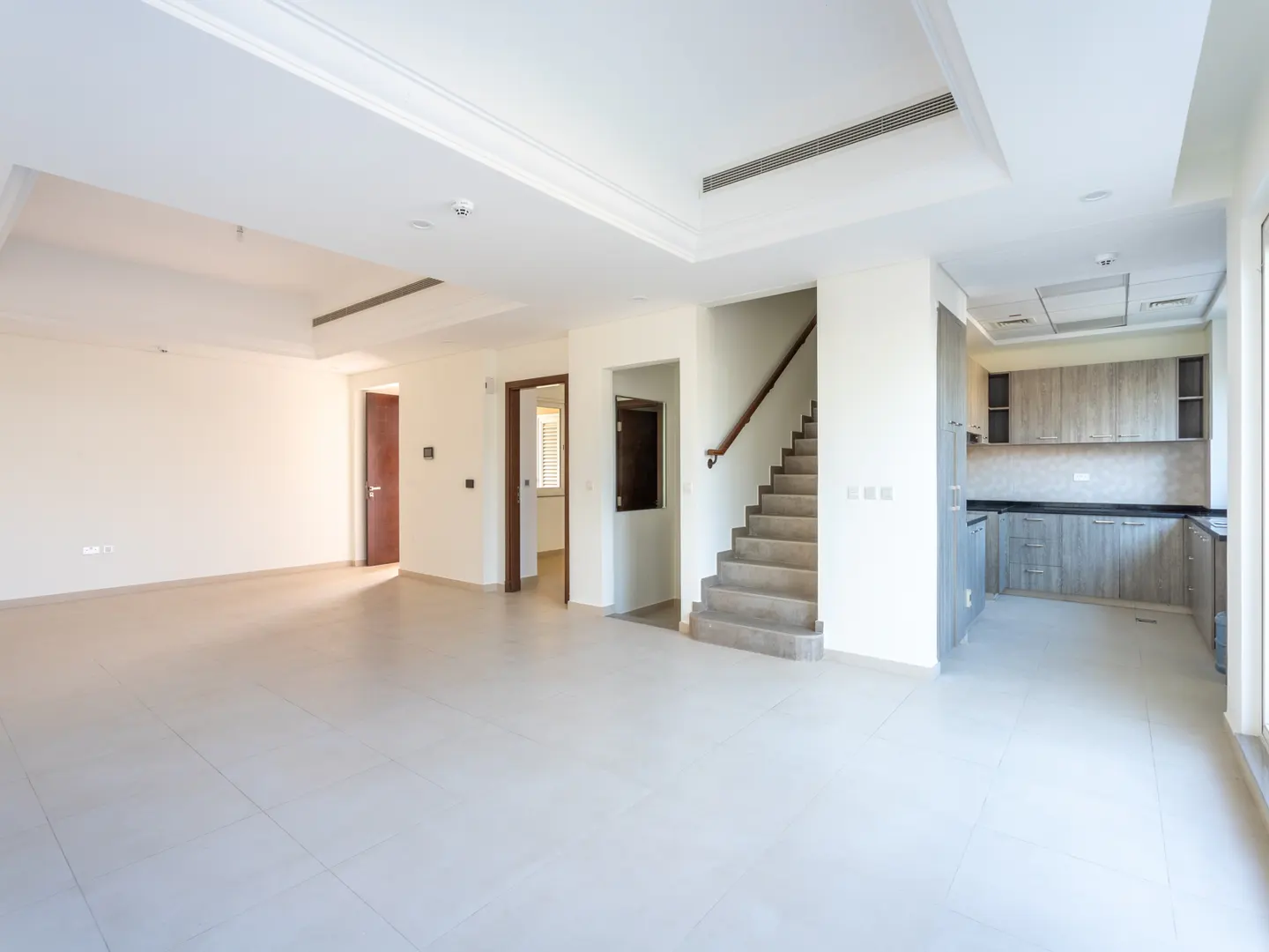 Bright, empty living room with tile floor, stairs, and kitchen with gray cabinets.