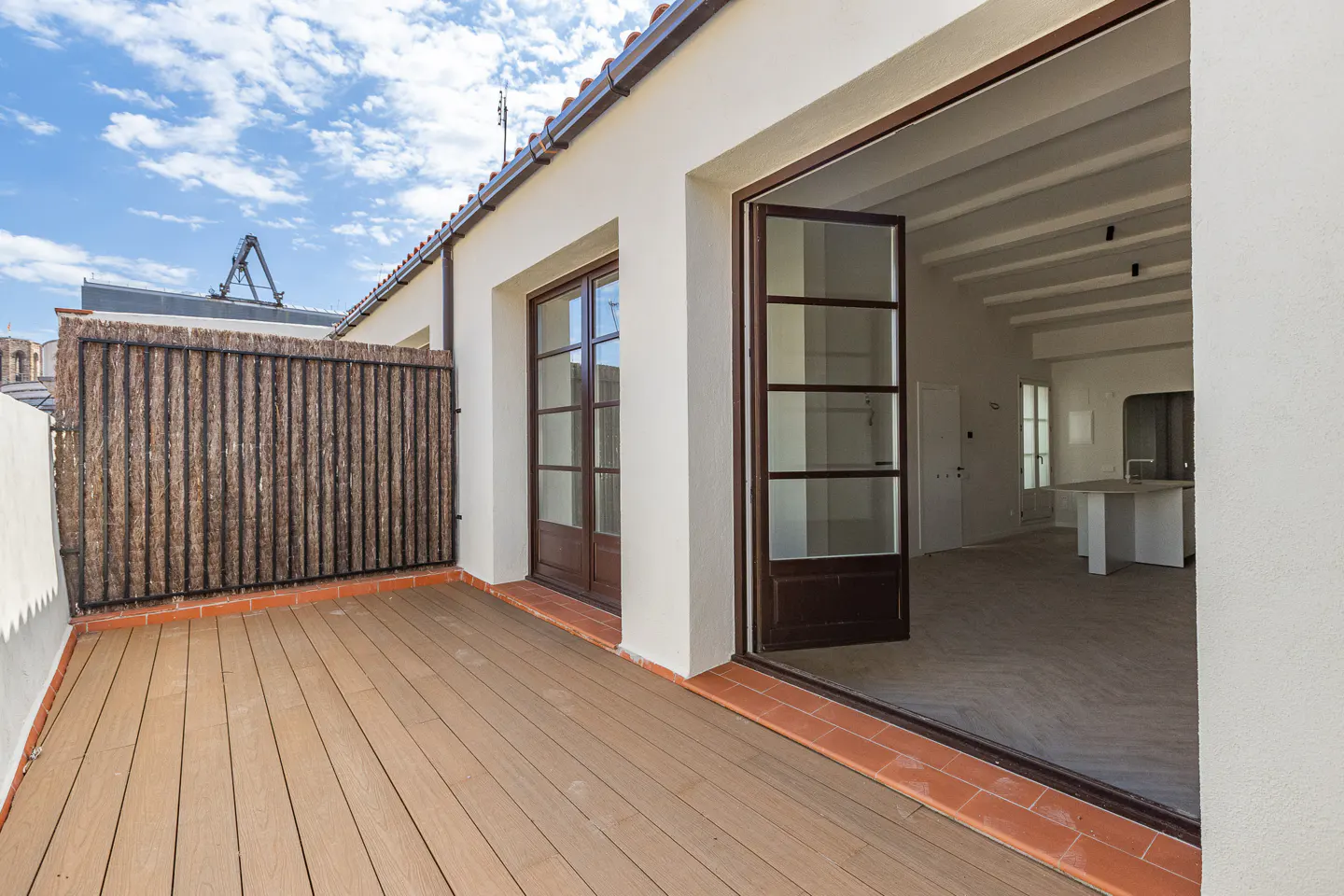 A bright, modern apartment balcony with wood flooring and a privacy screen. Open doors lead to a white interior with exposed beams.