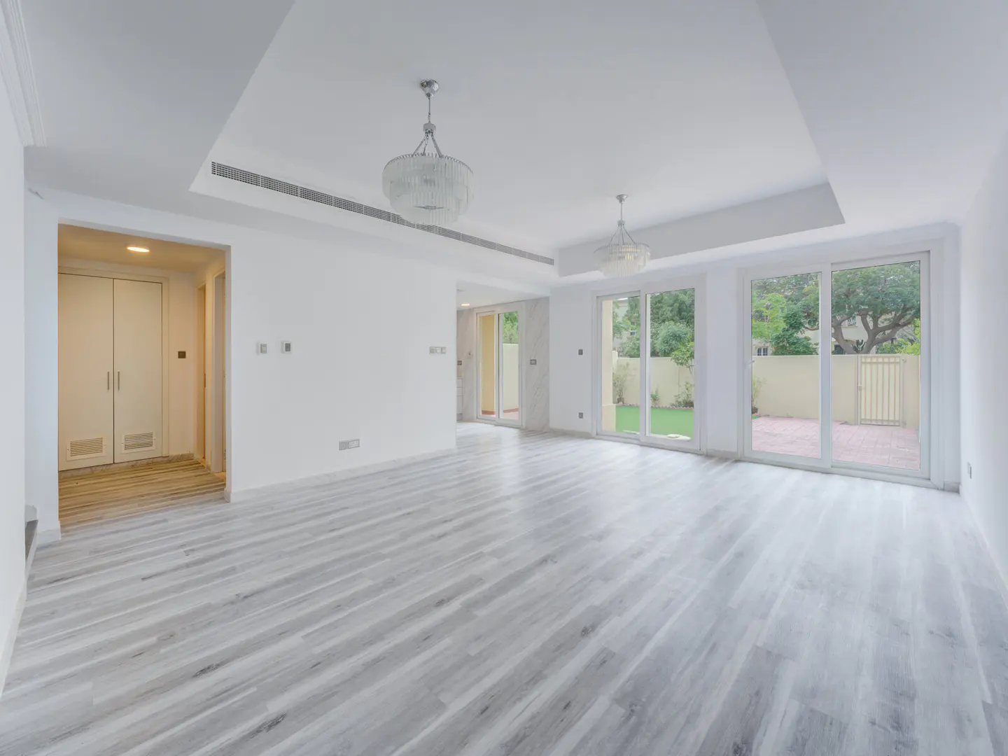 Bright, empty room with gray wood floors, white walls, and two chandeliers. Sliding glass doors lead to a green lawn and patio.