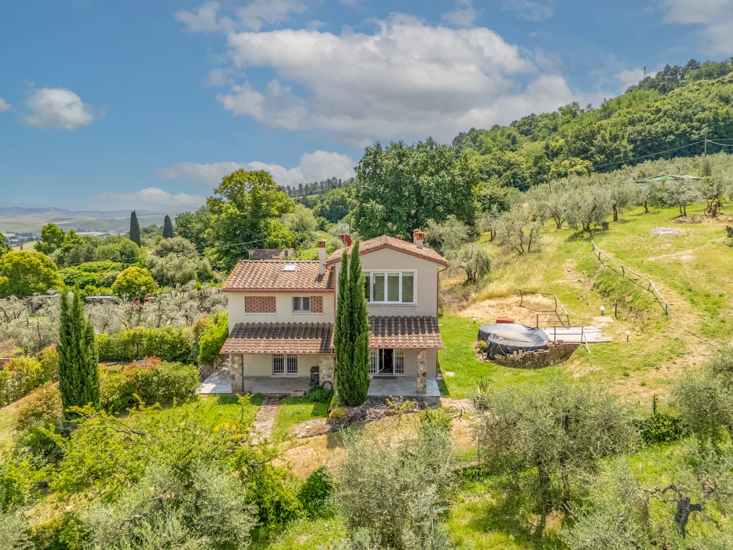 Two-story beige villa with a tiled roof, surrounded by olive trees and lush hills under a bright, cloudy blue sky.