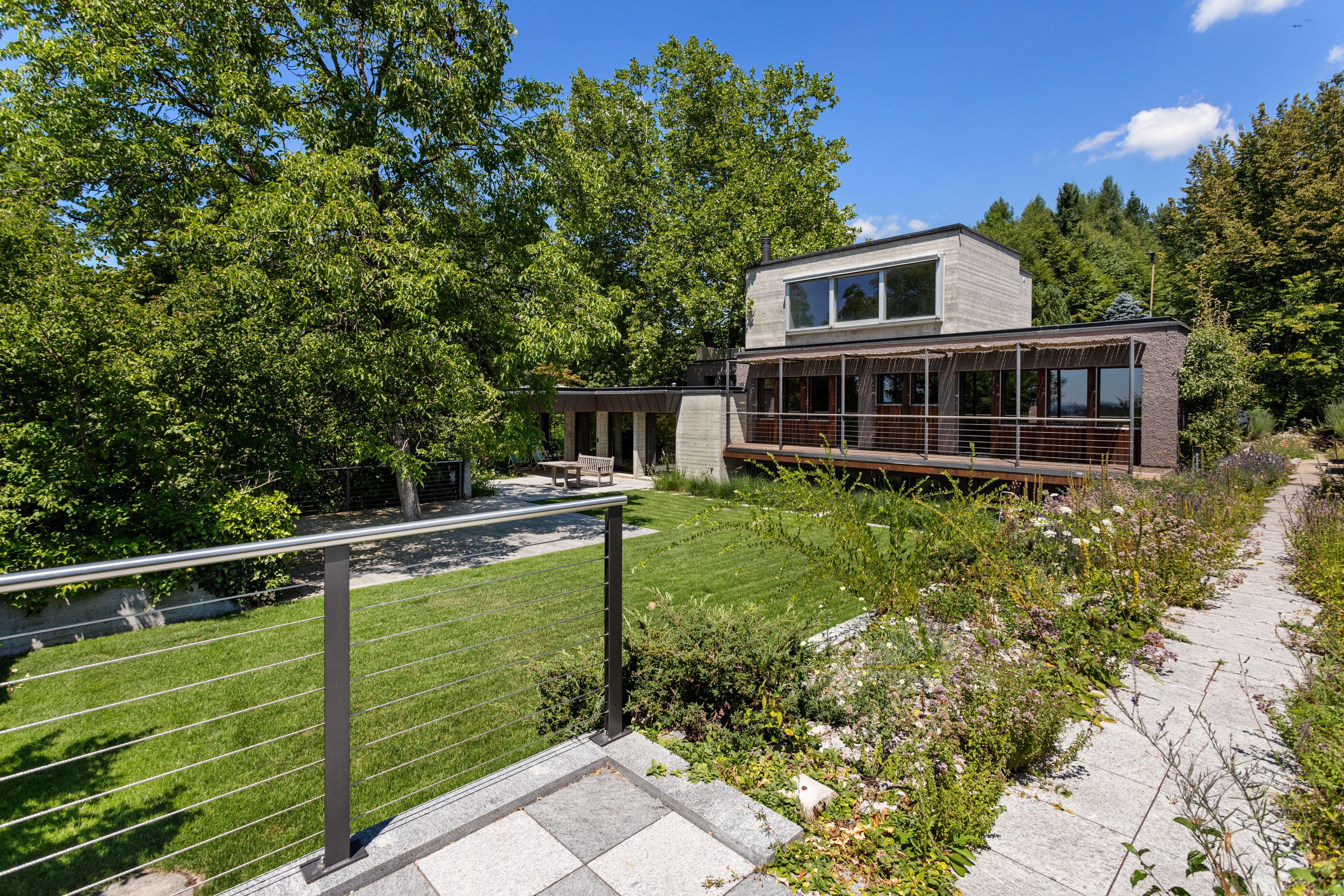 Modern two-story home with a wooden deck, lush green lawn, and stone path, surrounded by trees under a blue sky.