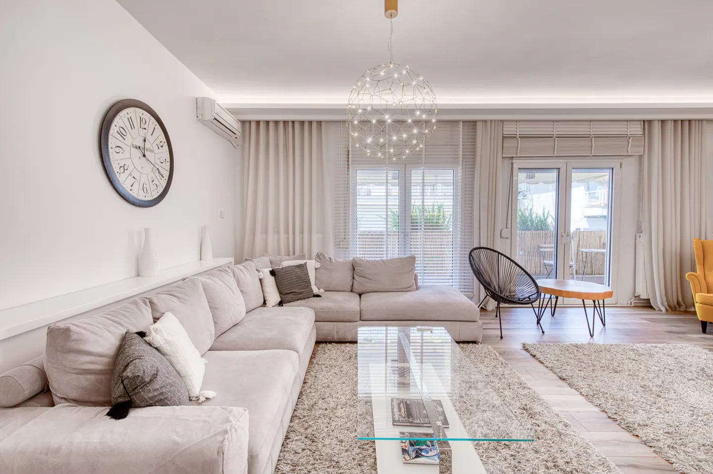 Modern living room with a large grey sectional sofa, glass coffee table, round wall clock, and a spherical pendant light.