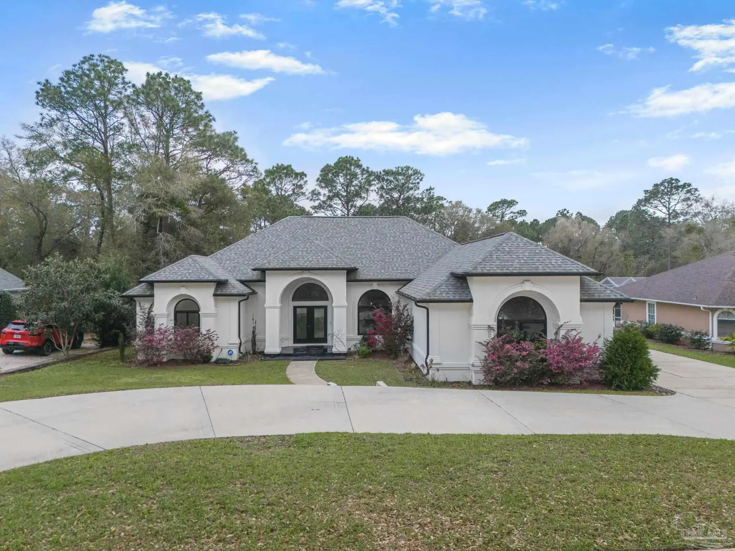 Single-story white stucco home with a gray shingled roof, arched windows, and a curved driveway under a blue sky.