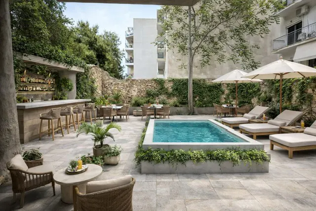 Outdoor patio with a pool, bar, and lounge chairs. The pool is surrounded by greenery. Tables and chairs are set for dining.