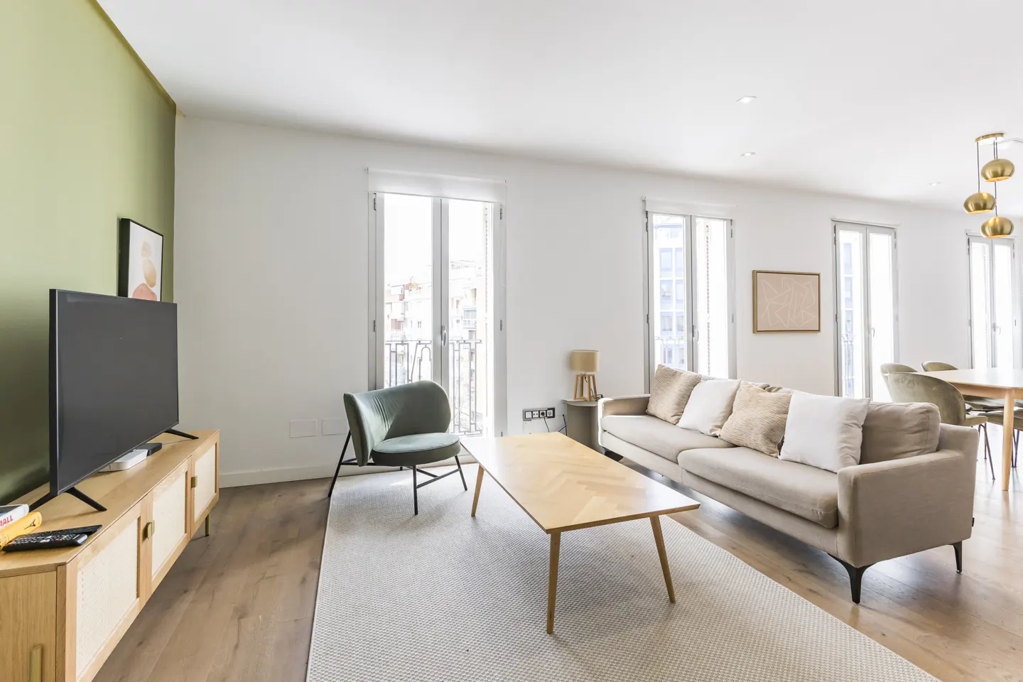 Bright living room with a beige sofa, wooden coffee table, green chair, and a TV on a wooden stand. Large windows let in natural light.