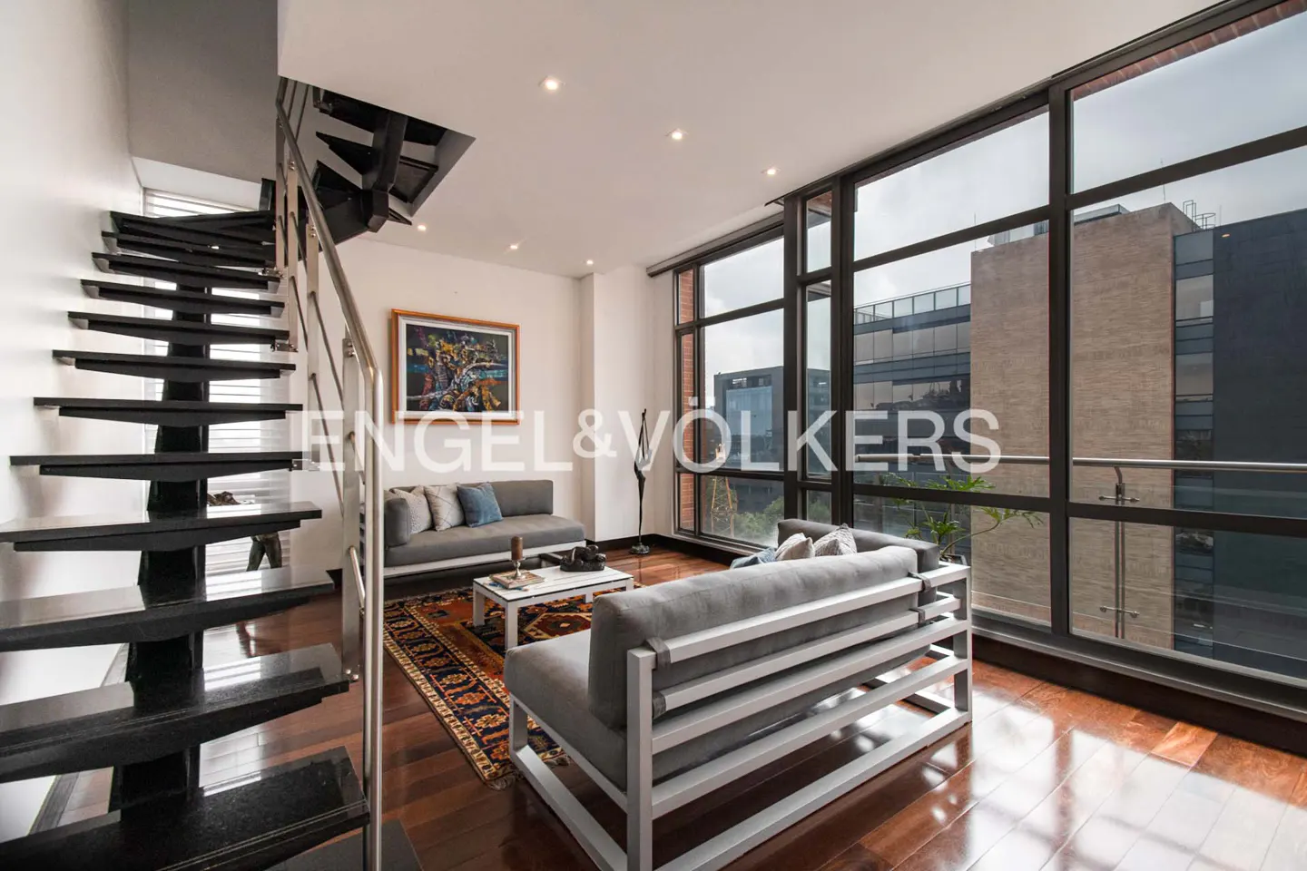 Living room with hardwood floors, gray sofas, and black stairs. Large windows overlook city buildings.