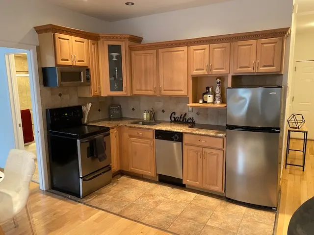 A kitchen with light wood cabinets, stainless steel appliances, and tan tile flooring. A "Gather" sign sits above the dishwasher.