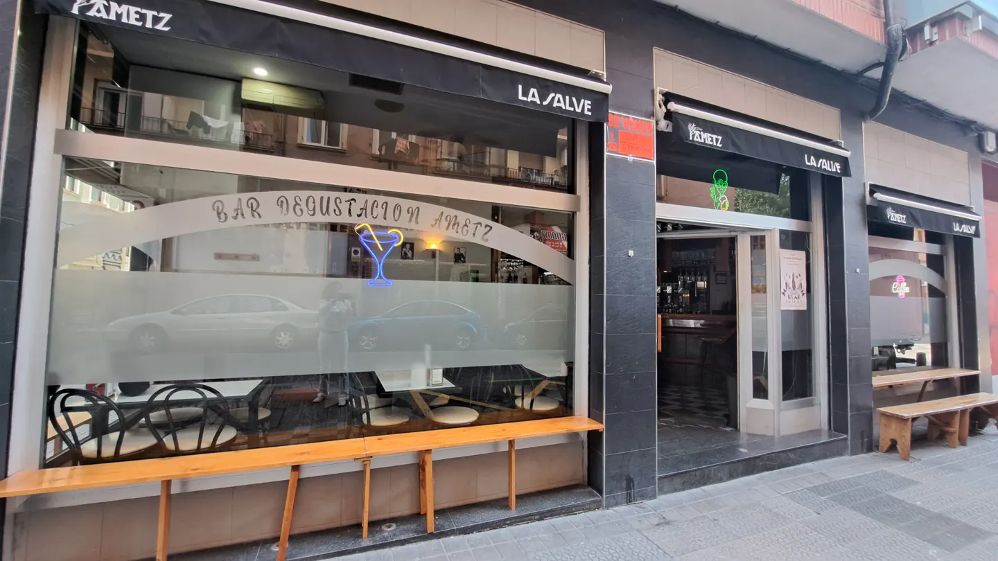 The exterior of "Bar Degustacion Ametz" with a large window, black awning, and wooden benches.
