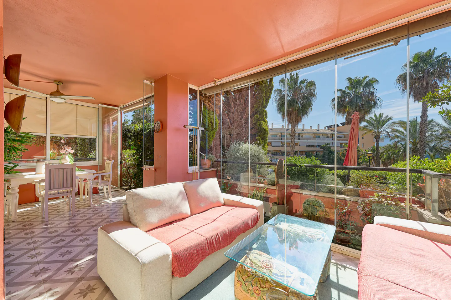 Bright sunroom with peach walls, tile floor, and glass walls. White sofas and a glass table sit in front of the glass walls with a view of palm trees. A white table and chairs are in the background.