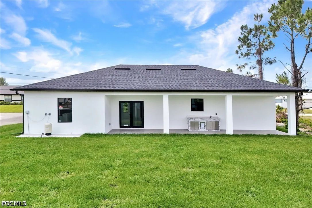 Back of a white house with a dark roof, black framed windows and doors, and a covered patio with an outdoor kitchen. Green lawn.