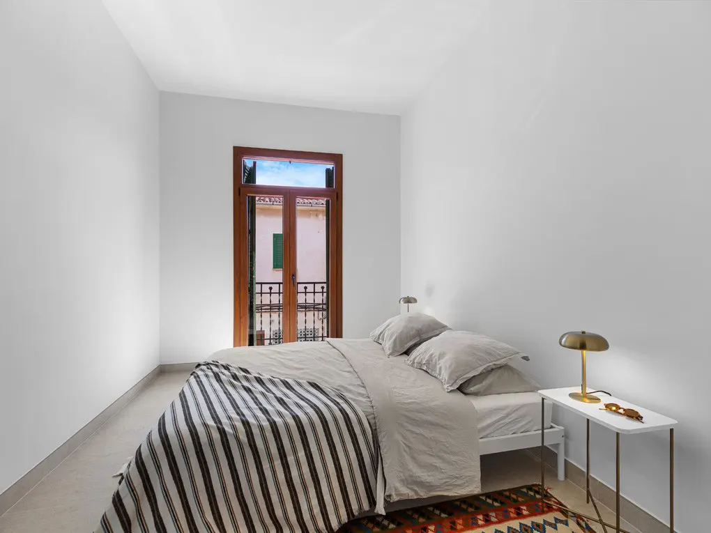 Bright bedroom with white walls, a bed with striped blanket, and a wooden door to a balcony. A small table with a lamp sits beside the bed.