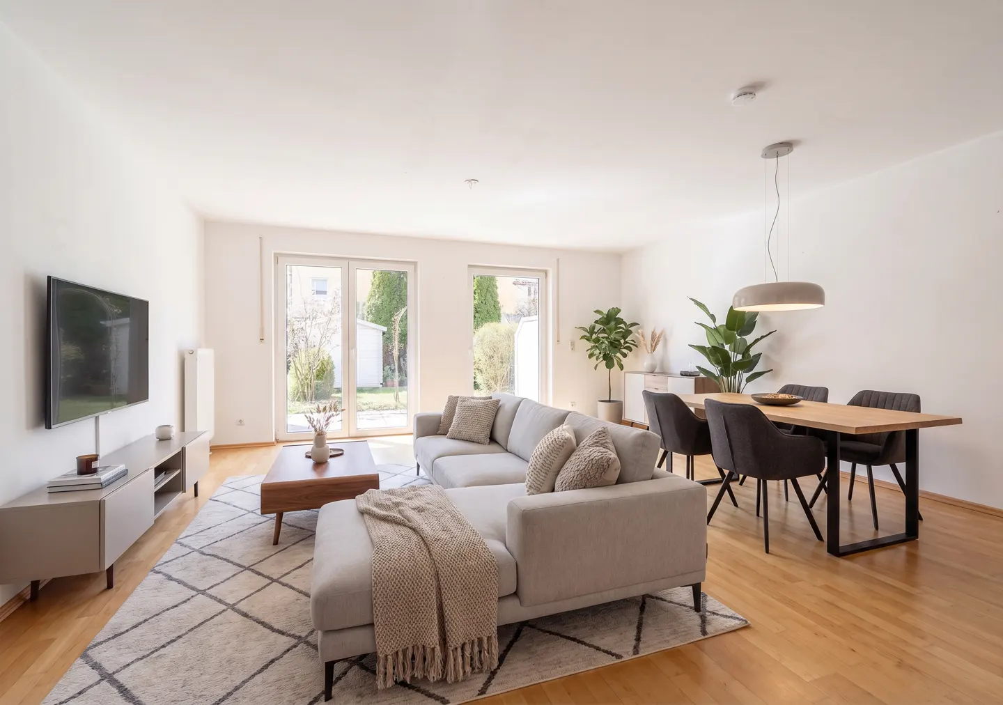 Bright living room with a gray sectional sofa, wood dining table with black chairs, and a TV. A patterned rug covers the wood floor.