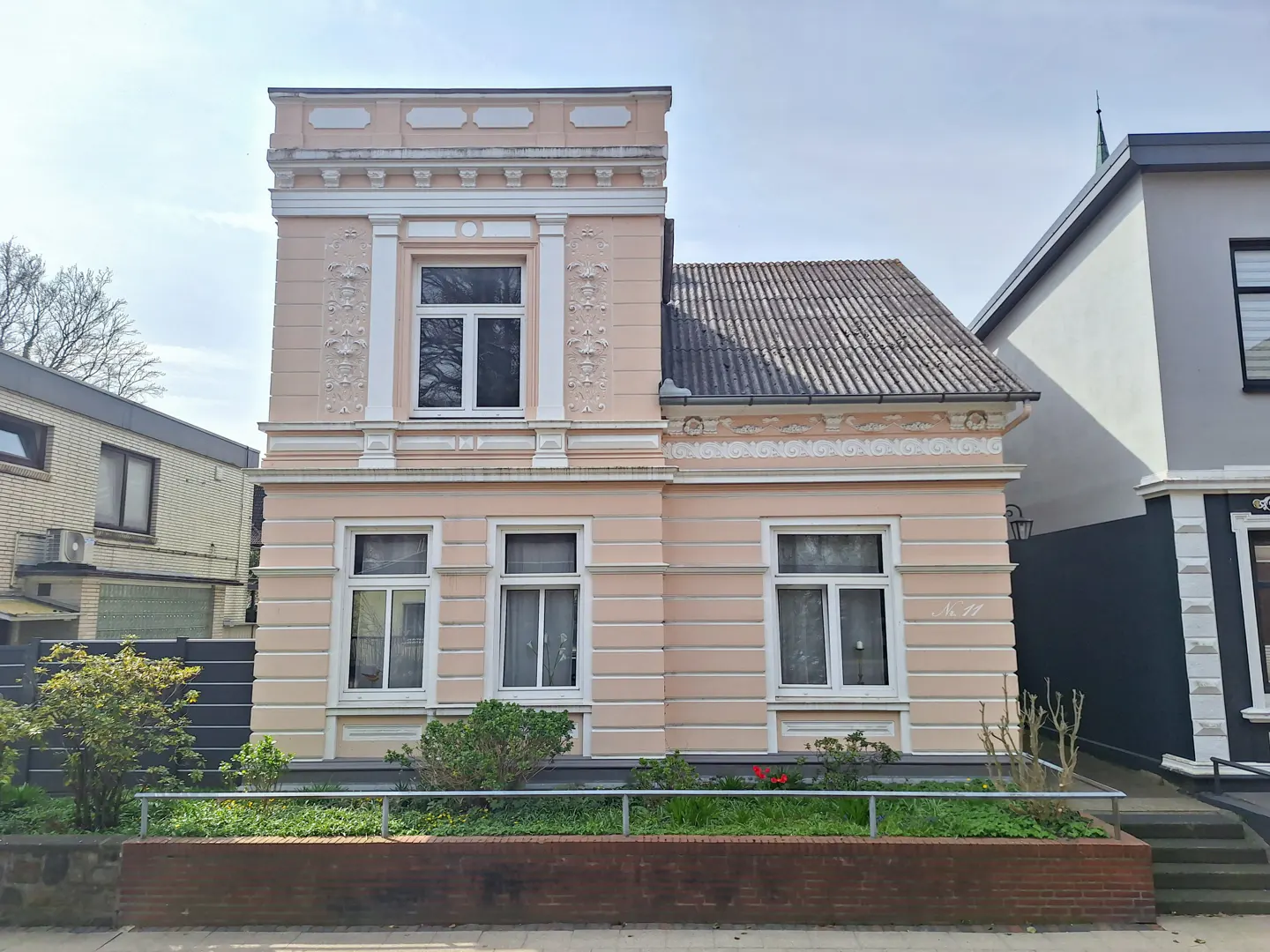 A two-story pink house with white trim and a gray roof. The house has a small garden in front.