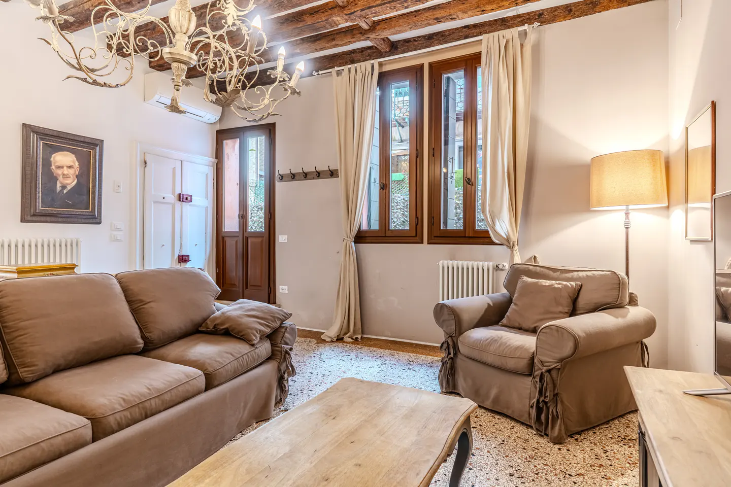 Cozy living room with brown sofa and armchair, wooden coffee table, ornate chandelier, and exposed ceiling beams.