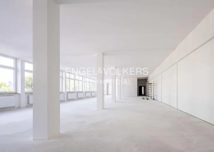 Bright, empty commercial space with white walls, pillars, and concrete floors. Large windows line one side, and a ladder stands in the back.