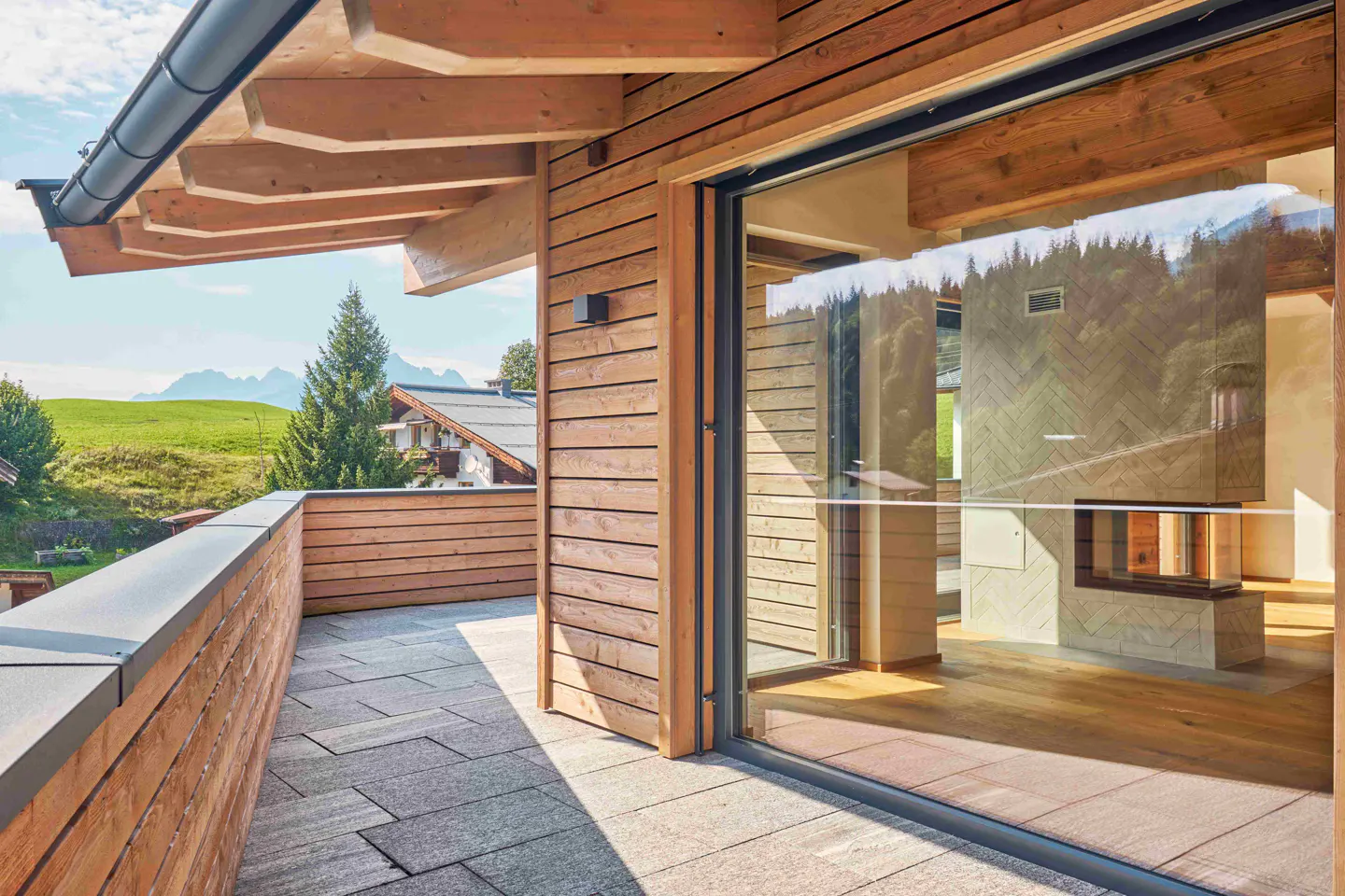 Balcony view of a wood-paneled home with a stone patio and sliding glass doors. A green field and mountains are visible in the background.