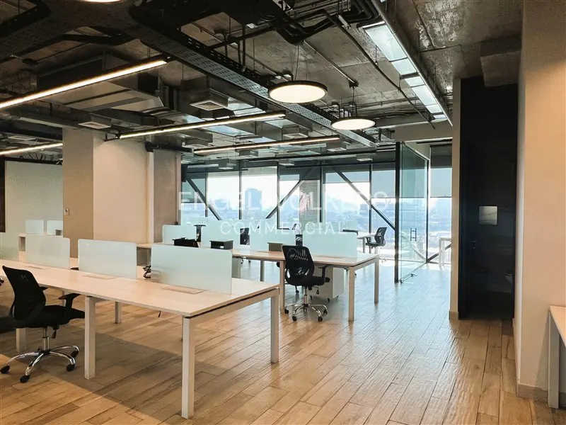 Bright, modern office space with desks, chairs, and large windows overlooking a city. The floor is light wood and the ceiling is exposed.