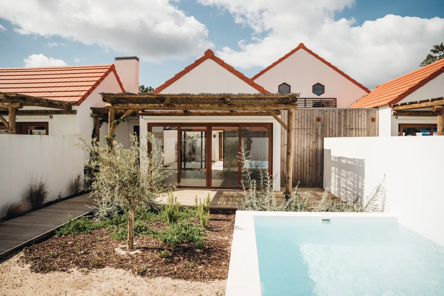 Exterior view of a white house with a red tile roof, a wooden pergola, and a blue swimming pool.
