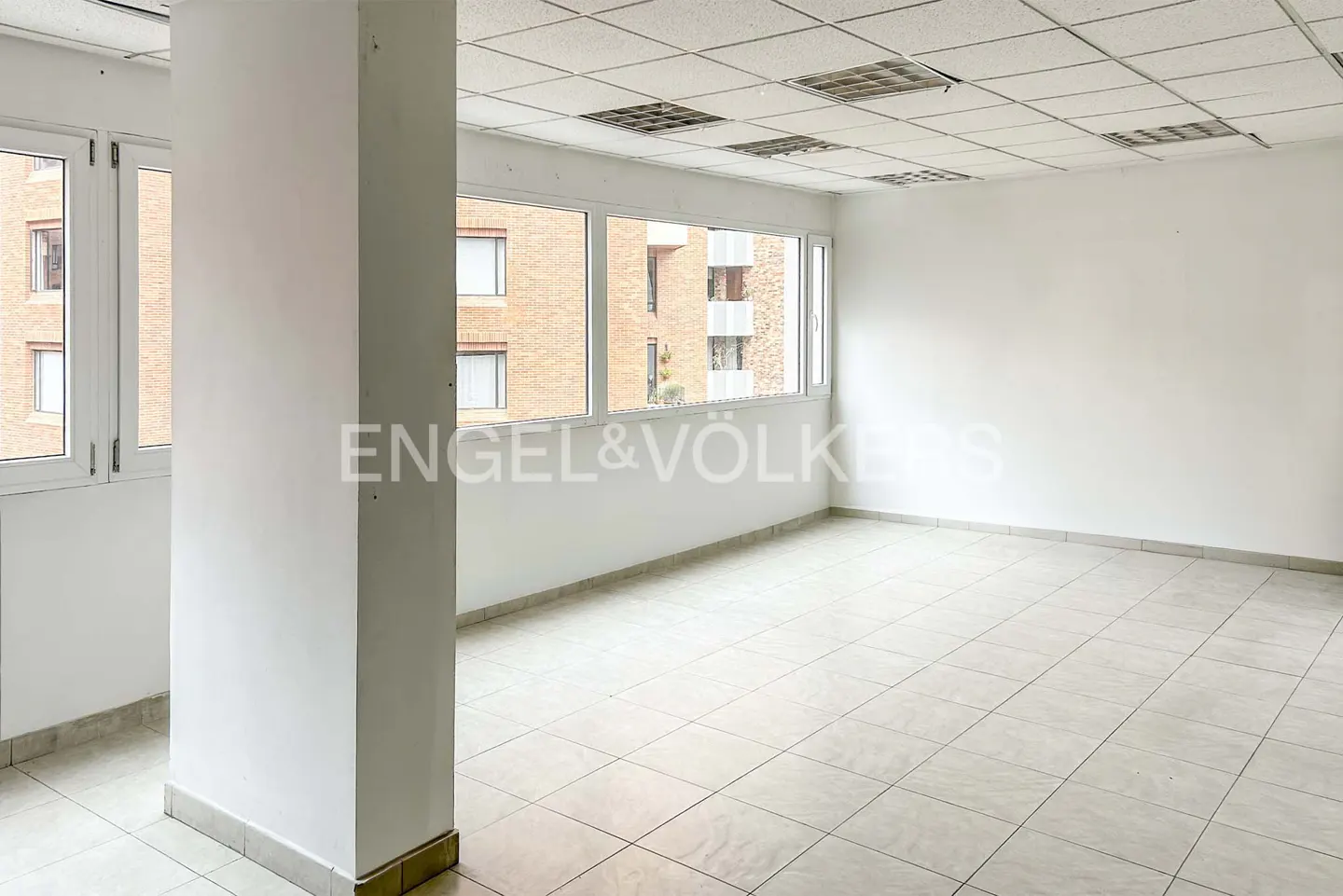 Bright, empty office space with white walls, tile floor, and large windows overlooking a brick building. Engel & Völkers logo visible.