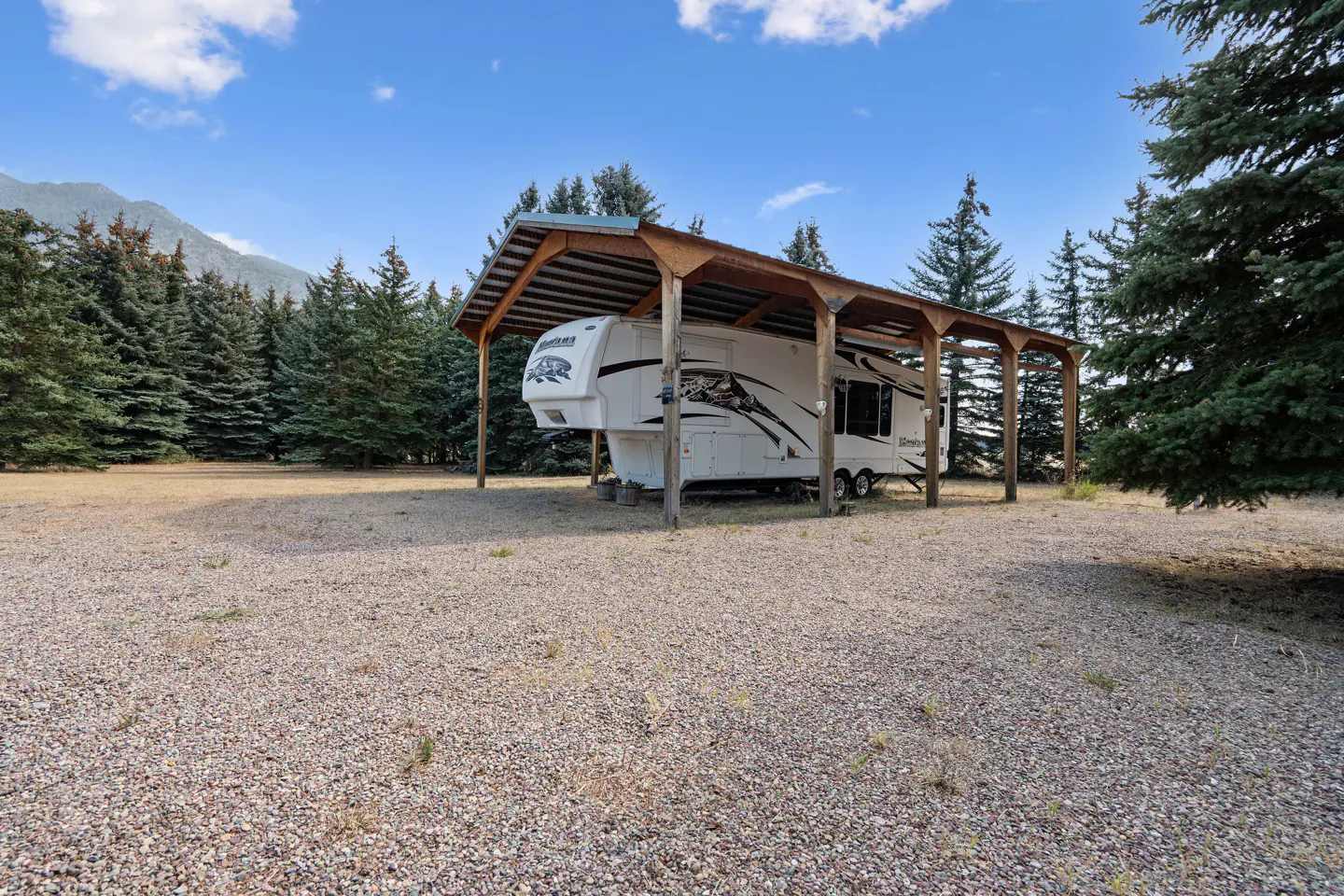 A white RV is parked under a wooden shelter on a gravel lot with pine trees and a mountain in the background.