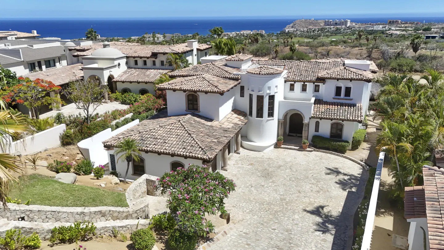 Aerial view of a white stucco house with a red tile roof, stone driveway, and ocean backdrop.