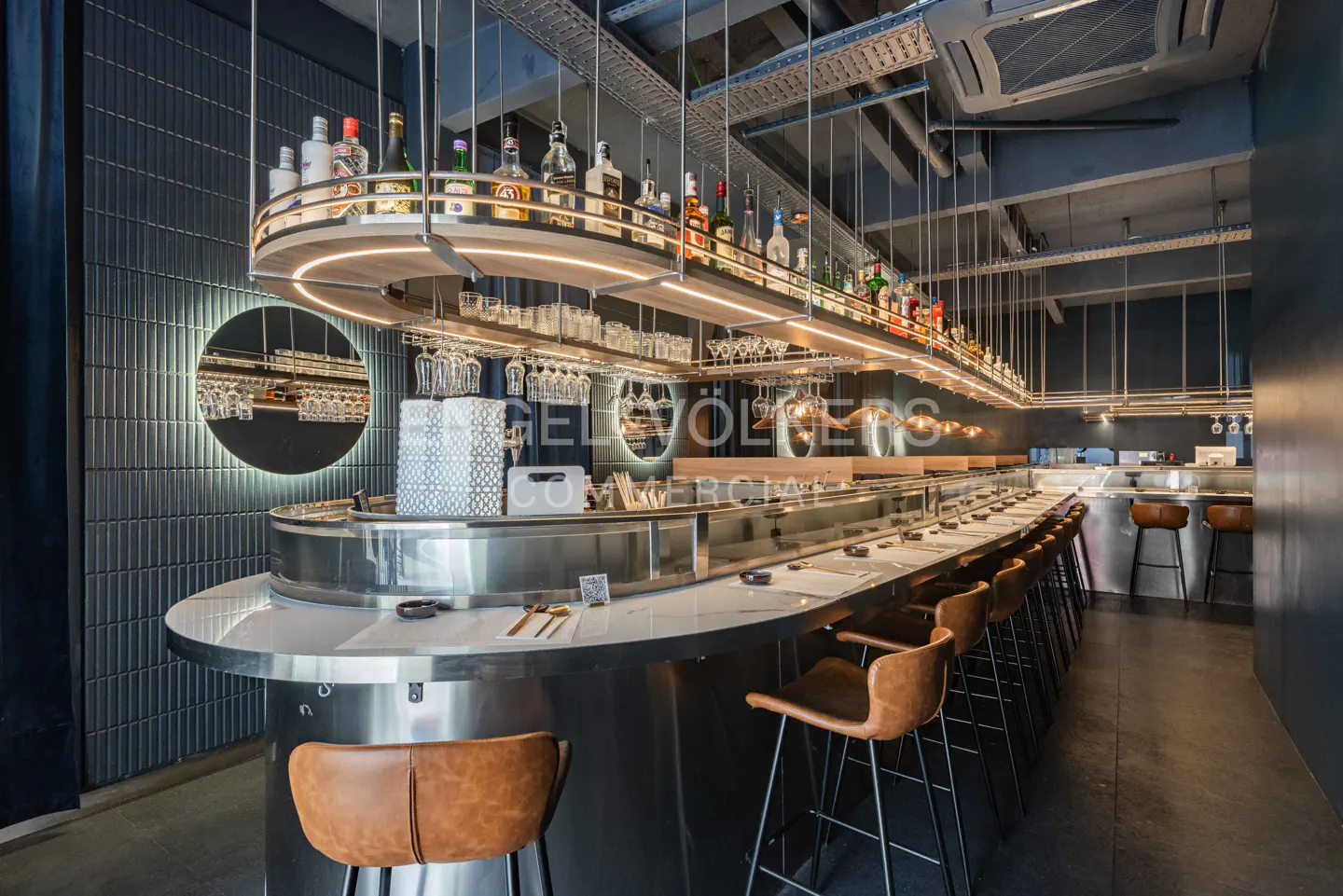 Interior view of a modern restaurant with a long, curved bar, brown leather stools, and a shelf of liquor bottles above.