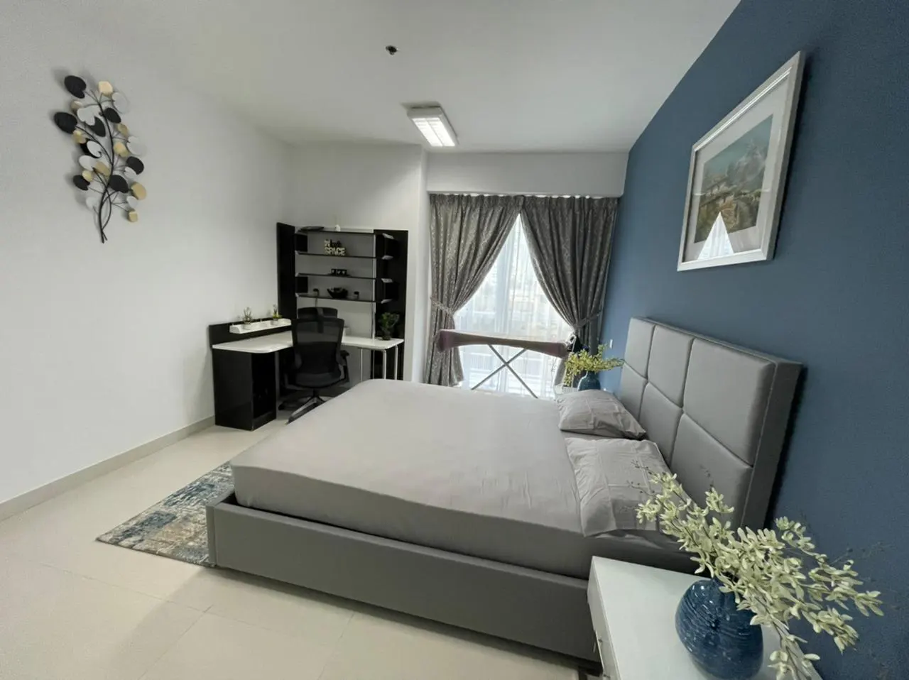 Bedroom with a gray bed, blue accent wall, desk, and shelves. A metal leaf sculpture hangs on the white wall. Curtains frame the window.