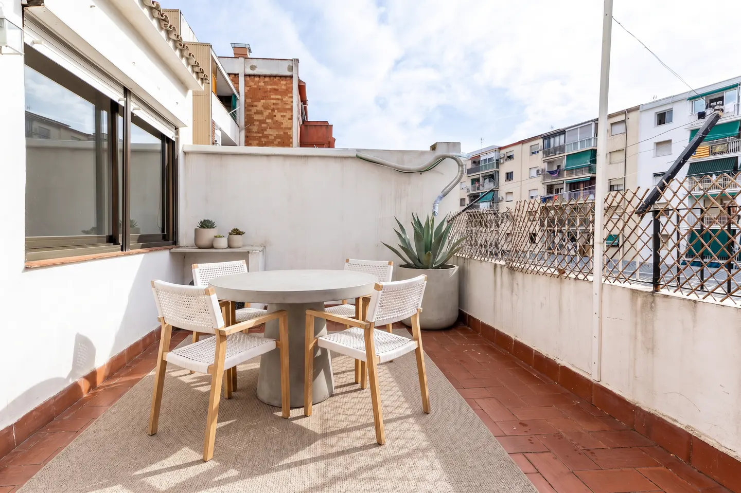 A bright outdoor patio with a round table and four chairs on a beige rug. A large potted plant sits near the white wall and fence.