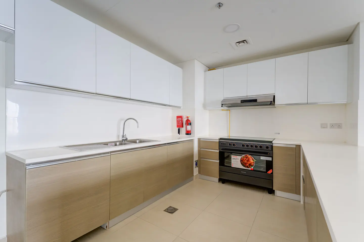 Bright kitchen with white cabinets, tan lower cabinets, stainless steel sink, and black oven. A fire extinguisher hangs on the wall.