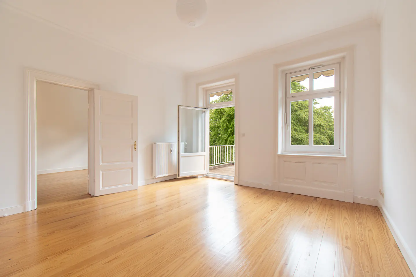 Bright, empty room with wood floors, white walls, and two windows. One window opens to a balcony with green trees visible. A door leads to another room.