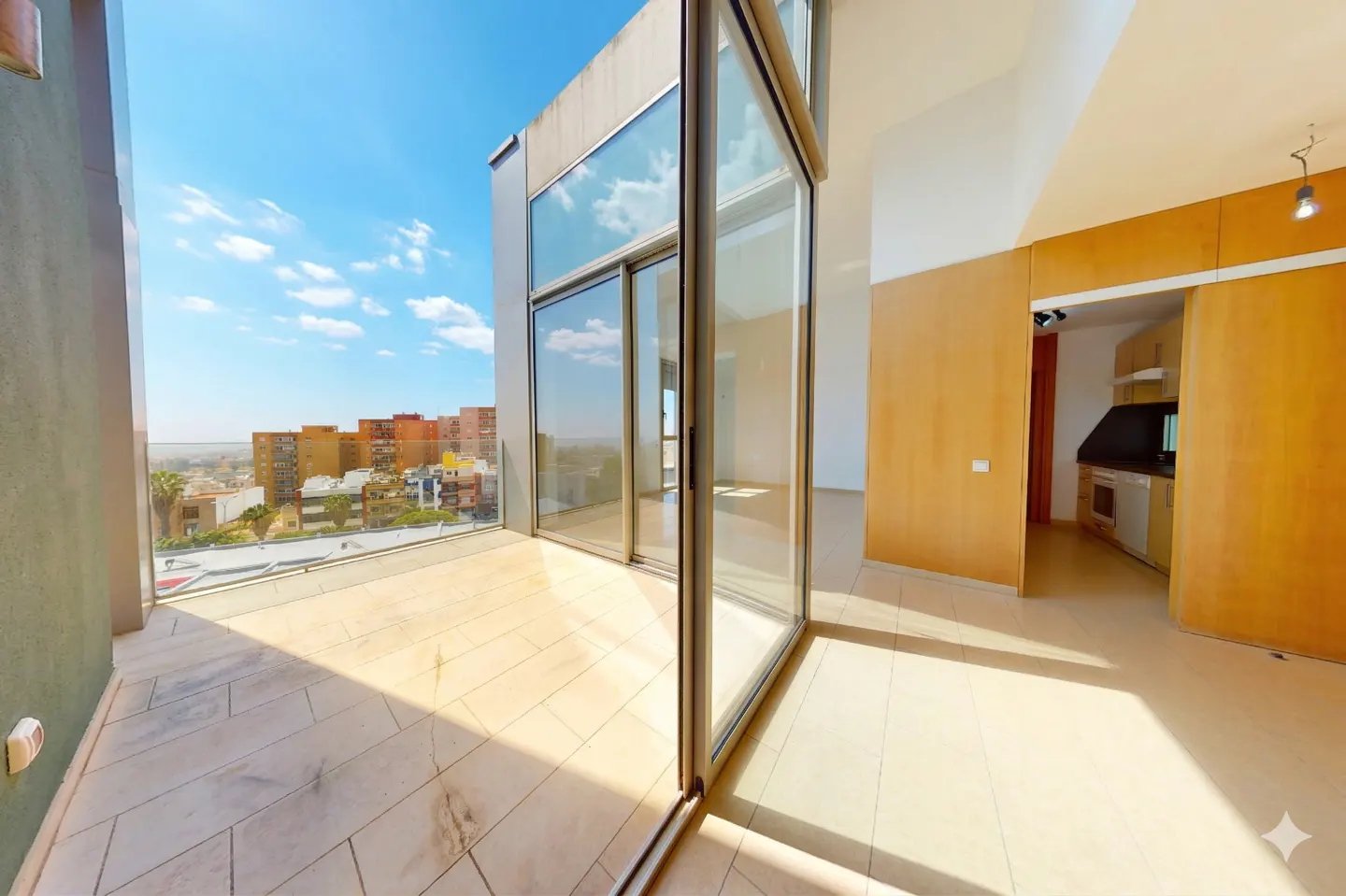 Modern apartment interior with a tiled balcony, floor-to-ceiling glass doors, and a view of city buildings under blue sky.