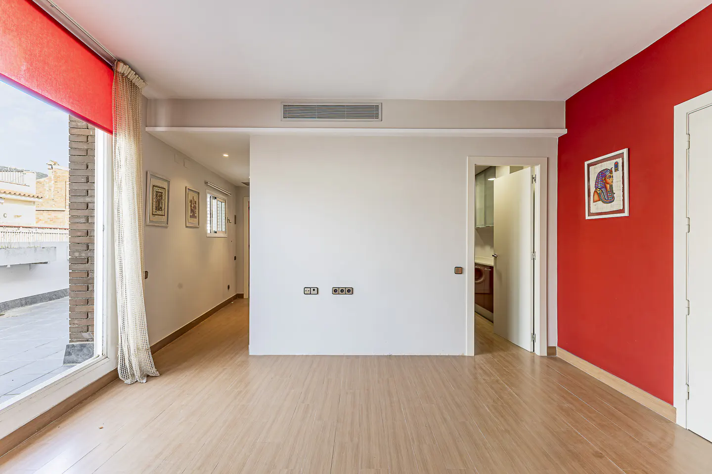 Bright, empty room with wood floors, white walls, and a red accent wall. A large window with a red shade offers a city view.