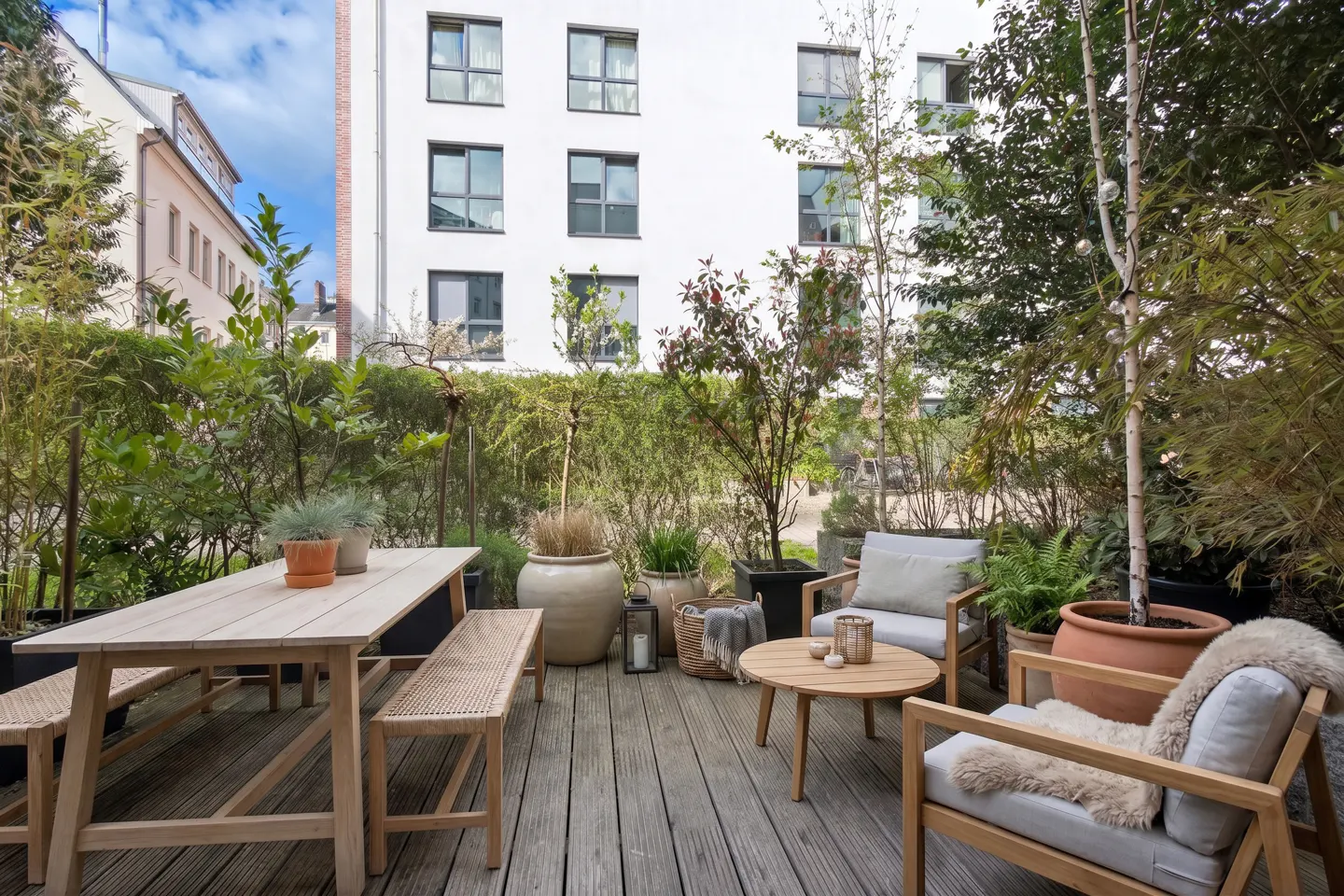 Outdoor patio with wooden furniture, including a table, benches, and chairs, surrounded by lush greenery and potted plants.