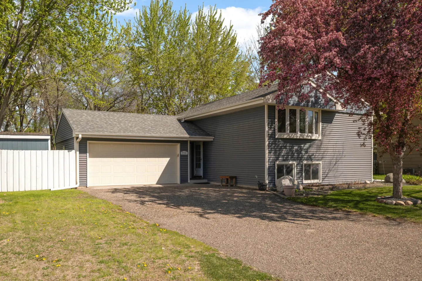 Two-story gray house with white garage door and gravel driveway. A pink-blossomed tree is on the right.
