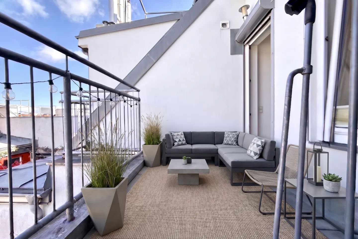 Rooftop patio with gray sectional sofa, coffee table, and potted plants on a woven rug. String lights hang from the black metal railing.