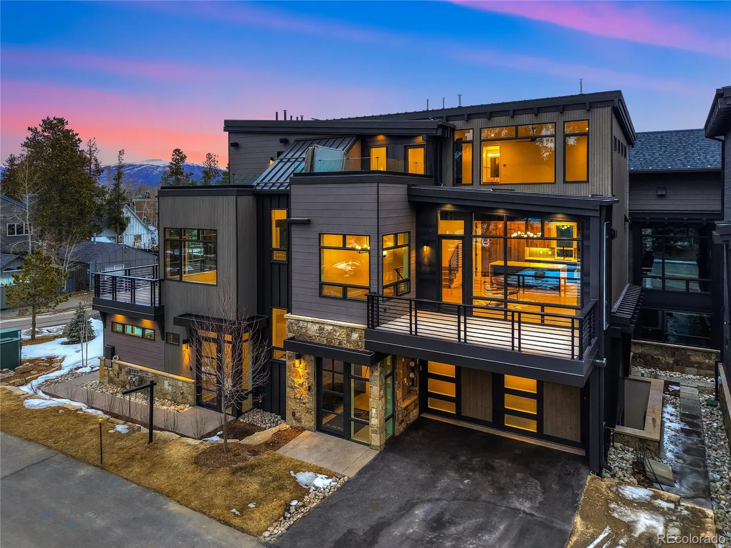 Exterior view of a modern, multi-story home with gray siding, black trim, and large windows at dusk. Balconies and a driveway are visible.