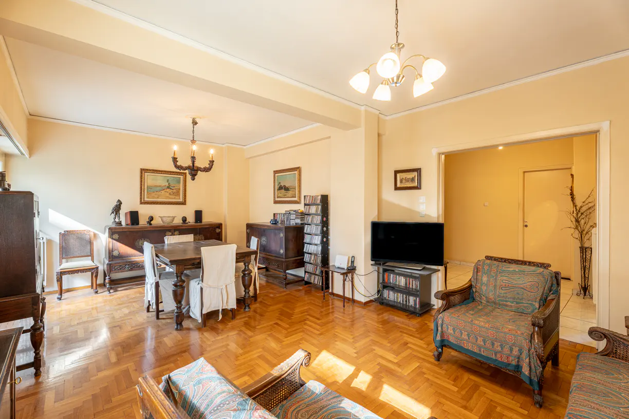 A bright living room with a dining area, wooden floors, and antique furniture. A TV and patterned armchairs are visible.