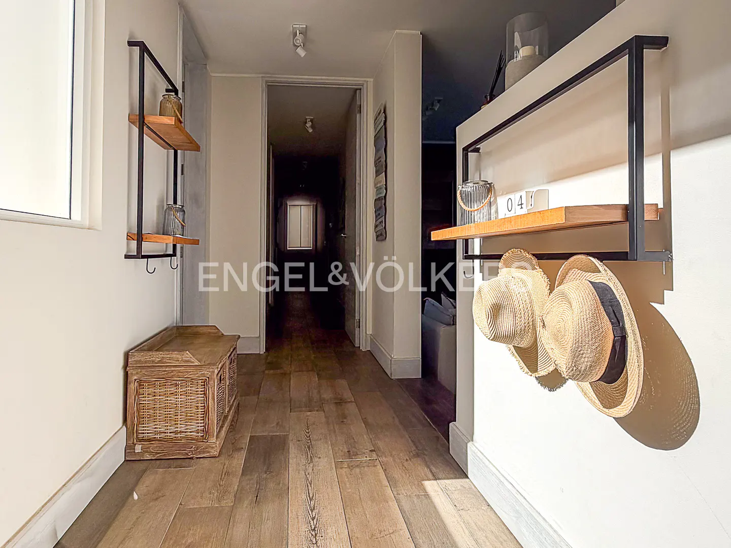 Hallway with light wood floors, white walls, and a dark doorway. A wooden bench sits near a black shelf with hats.