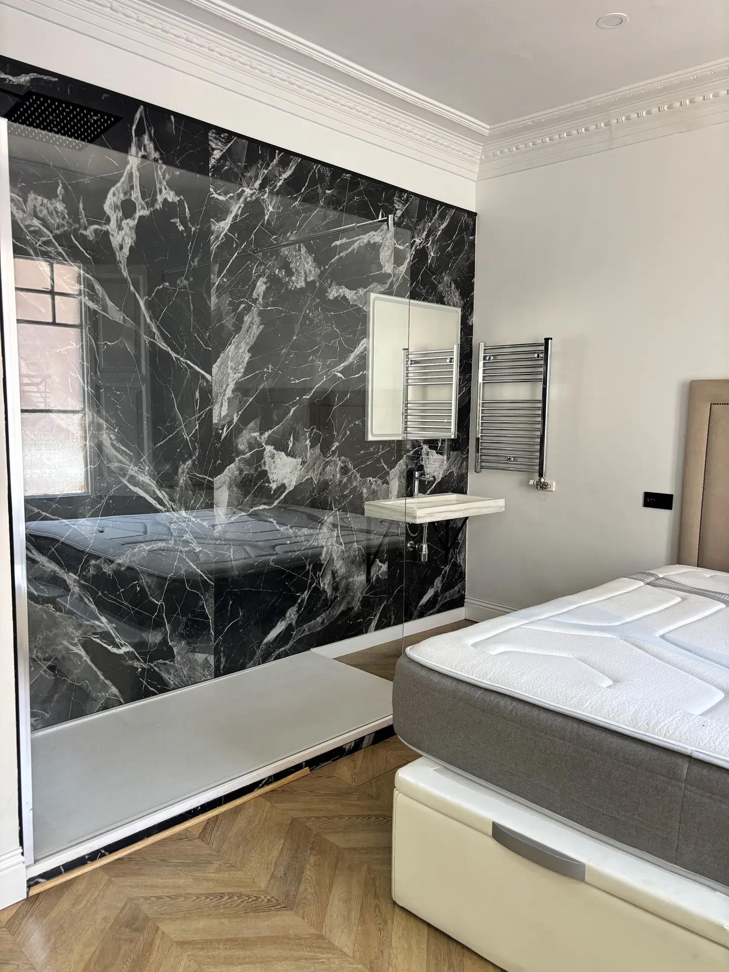 Bedroom with a black marble shower, white sink, and heated towel rack. A bed with a white mattress is in the foreground.