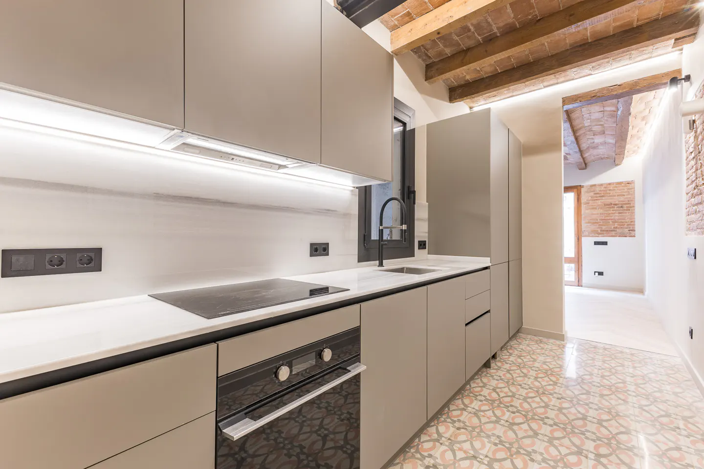 Modern kitchen with gray cabinets, white countertops, black appliances, and patterned tile floor. Exposed brick ceiling beams add rustic charm.